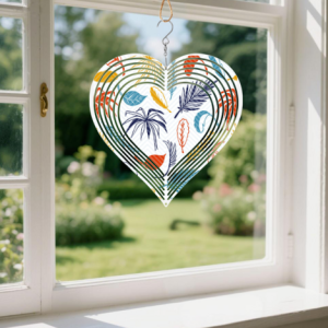 Custom heart-shaped wind spinner with a colorful leaf print, hanging in a window with a garden view in the background.