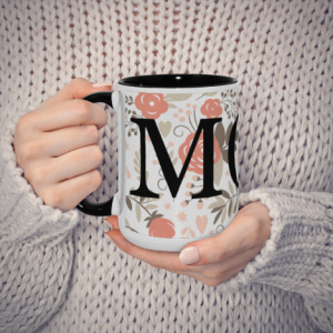 Hands holding a white ceramic mug with a black interior and handle, featuring a large "M" monogram with pink floral and heart patterns, perfect as a Mother’s Day gift.