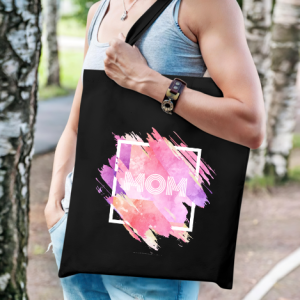 Black canvas tote bag with a pink watercolor “MOM” print, worn by a woman outdoors, ideal as a practical Mother’s Day gift.