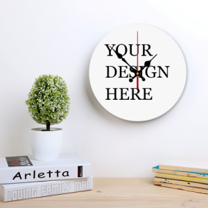 Custom round wall clock mockup with "YOUR DESIGN HERE" text, hung on a light-colored wall above a wooden desk with books and a potted plant, showing office or workspace use.