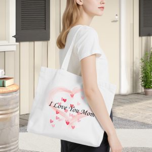 Woman carrying a white canvas tote bag with a pink heart print and "I Love You Mom" text, perfect as a Mother’s Day gift.