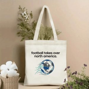 Beige canvas tote bag featuring "football takes over north america" text and a stylized globe with North America map print, displayed with cotton branches and dried botanicals, fan accessory