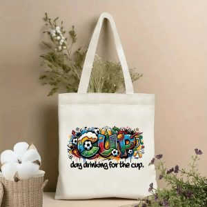 Front view of beige canvas tote bag with vibrant graffiti-style "CUP" lettering filled with soccer balls, beer mugs, and stadium motifs. Below is the slogan "day drinking for the cup." Styled with cotton and dried botanicals, 2026 World Cup game day accessory.