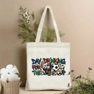 Front view of beige canvas tote bag featuring colorful graffiti-style slogan "Day Drinking For The Cup" with a cartoon fan holding a trophy, soccer ball, and beer mug. Styled with cotton and dried botanicals, 2026 World Cup game day accessory.