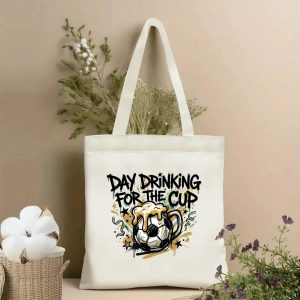 Front view of beige canvas tote bag with bold graffiti-style "Day Drinking For The Cup" slogan, featuring a creative graphic of a soccer ball merged with a frothy beer mug. Styled with cotton and dried botanicals, 2026 World Cup game day accessory.