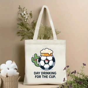 Front view of beige canvas tote bag with a playful graphic of a soccer beer mug, a cactus with a Canadian maple leaf, and the slogan "DAY DRINKING FOR THE CUP." Styled with cotton and dried botanicals, 2026 World Cup game day accessory.