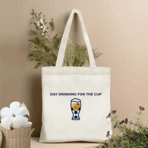 Front view of beige canvas tote bag with bold navy text "DAY DRINKING FOR THE CUP" above a stylized soccer beer pint glass graphic featuring a sun icon. Styled with cotton and dried botanicals, 2026 World Cup game day accessory.
