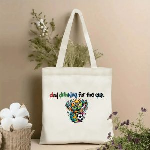 Front view of a beige canvas tote bag showcasing a vibrant graffiti-style print. The design features colorful, dripping lettering spelling "day drinking for the cup" above a chaotic street-art graphic of beer bottles, a soccer ball, kegs, and the word "BREW" with sun rays and star accents. Styled with cotton stems and dried botanicals in the background.