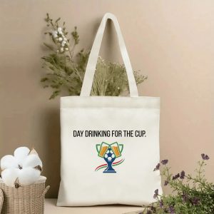 Front view of a beige canvas tote bag with the slogan "DAY DRINKING FOR THE CUP" in bold black font. The design includes a central graphic of a soccer ball trophy with two clinking beer mugs and colorful accent details. Styled against a backdrop of cotton stems and dried botanicals.
