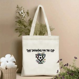 Front view of a beige canvas tote bag with bold graffiti-style "DAY DRINKING FOR THE CUP" lettering, paired with a vibrant hand-drawn graphic of a soccer ball trophy, beer splashes, a bottle, and a sun icon. Styled with cotton stems and dried botanicals, perfect for 2026 World Cup game days.