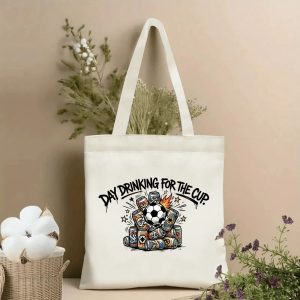 Front view of a beige canvas tote bag with a playful 2026 World Cup-themed print. The design includes dripping graffiti lettering "DAY DRINKING FOR THE CUP" and a dynamic graphic of a soccer ball surrounded by stacked, colorful beer cans with flame and star accents, styled with cotton stems and dried botanicals.