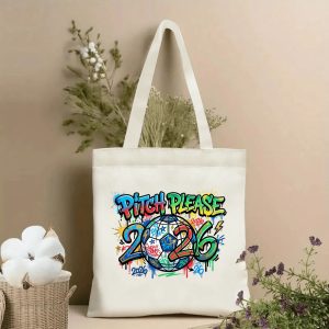 Front view of a beige canvas tote bag with a vibrant 2026 World Cup graffiti-style print, featuring bold "PITCH PLEASE" lettering, a colorful soccer ball, and "2026" numerals with paint splatters and street art details, styled with cotton stems and dried botanicals.