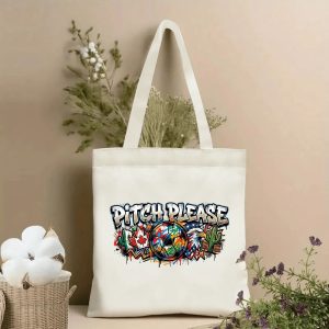 Front view of a beige canvas tote bag with a vibrant 2026 World Cup graffiti-style print, featuring bold "PITCH PLEASE" lettering, a colorful soccer ball, Canadian maple leaf, bald eagle, and cactus motifs with paint splatter details, styled with cotton stems and dried botanicals.