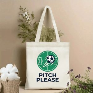 Front view of a beige canvas tote bag with a minimalist 2026 World Cup-themed print, featuring a green circular soccer pitch graphic, a dynamic 3D soccer ball, and bold navy "PITCH PLEASE" lettering, styled with cotton stems and dried botanicals.