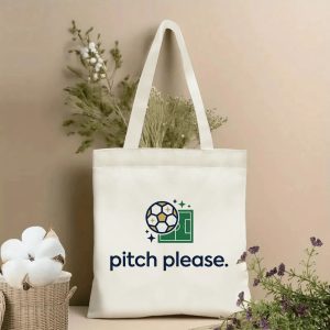 Front view of a beige canvas tote bag with a whimsical 2026 World Cup print, featuring a stylized soccer ball with star accents, a green pitch graphic, and the playful lowercase slogan "pitch please.", styled with cotton stems and roses.