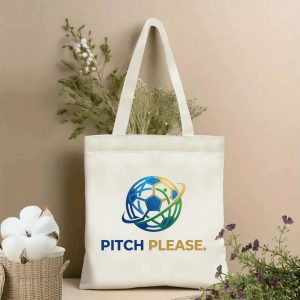 Front view of a beige canvas tote bag featuring a vibrant 2026 World Cup graphic logo. The design showcases a stylized soccer ball with interconnected blue and green lines, accented with a golden orbital ring, paired with the bold split slogan "PITCH PLEASE." (PITCH in blue, PLEASE. in gold). Styled with cotton stems and roses in the background.