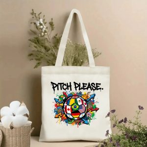 Front view of a beige canvas tote bag with a vibrant 2026 World Cup graffiti print, featuring bold dripping "PITCH PLEASE." lettering, a multicolored soccer ball decorated with international flags, and street art splatters, arrows, and cultural motifs, styled with cotton stems and dried botanicals.
