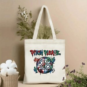 Front view of a beige canvas tote bag with a vibrant 2026 World Cup graffiti print. Features the slogan "PITCH PLEASE." in red, green and blue drip lettering, a stylized soccer ball with "26" graphic, Canadian maple leaf and Mexican cactus motifs, styled with cotton stems and roses.