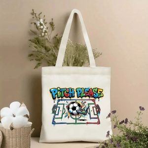 Front view of a beige canvas tote bag with a vibrant 2026 World Cup graffiti print. Features the bold "PITCH PLEASE" slogan in multicolored drip lettering, a stylized soccer pitch graphic with a kicking foot and soccer ball, surrounded by paint splatters, styled with cotton stems and dried botanicals.