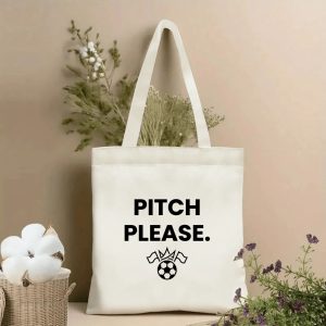 Front view of a beige canvas tote bag with a minimalist 2026 World Cup design. Features the bold black sans-serif slogan "PITCH PLEASE." stacked above a simple black line art icon of a soccer ball with a crown and two flags, styled with cotton stems and roses.