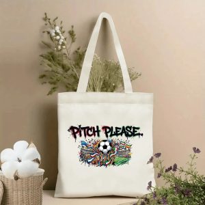 Front view of a beige canvas tote bag with a vibrant 2026 World Cup graffiti print. Features the bold dripping "PITCH PLEASE." lettering, a central soccer ball held aloft by cheering hands, international flags, and a stadium scene, styled with cotton stems and dried botanicals.