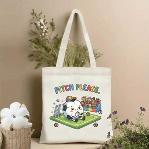 Front view of a beige canvas tote bag with a cute 2026 World Cup kawaii design. Features the playful "PITCH PLEASE." slogan in rainbow lettering, a chibi-style anthropomorphic soccer ball character in a cap sitting on a pitch, with a goal, cheering kids, stars, and mini soccer balls, styled with cotton stems and dried botanicals.