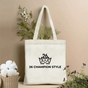 Front view of a beige canvas tote bag with a minimalist 2026 World Cup design. Features a sleek black line-art logo of a crowned soccer ball with "26" detail, paired with the bold "26 CHAMPION STYLE" text, styled with cotton stems and dried botanicals.