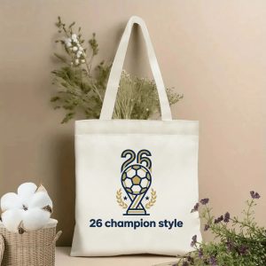 Front view of a beige canvas tote bag with a 2026 World Cup champion design. Features a trophy-style logo with a soccer ball, number "26", laurel wreaths, stars, and the "26 champion style" slogan, styled with cotton stems and dried botanicals.