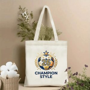 Front view of a beige canvas tote bag with a 2026 World Cup champion design. Features a circular emblem with a stylized soccer ball, number "26", laurel wreaths, stars, radiant lines, and the "CHAMPION STYLE" slogan, styled with cotton stems and dried botanicals.
