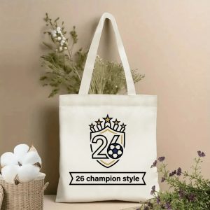 Front view of a beige canvas tote bag with a 2026 World Cup champion design. Features a shield-style emblem with the number "26", a soccer ball, five stars, and the "26 champion style" banner slogan, styled with cotton stems and dried botanicals.