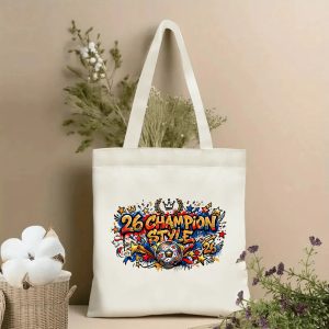 Front view of a beige canvas tote bag with a vibrant 2026 World Cup graffiti design. Features bold "26 CHAMPION STYLE" lettering, a stylized soccer ball, crown, laurel wreath, stars, and dynamic street-art style details, styled with cotton stems and dried botanicals.