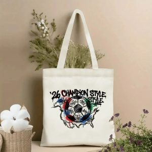 Front view of a beige canvas tote bag with a bold 2026 World Cup street art design. Features graffiti-style "'26 CHAMPION STYLE" lettering, a hand-drawn soccer ball with tag details, and a map outline with spray-paint drips, styled with cotton stems and dried botanicals.