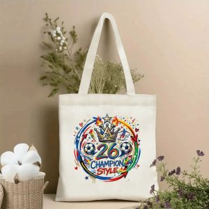 Front view of a beige canvas tote bag with a vibrant 2026 World Cup celebration design. Features a circular graphic with a crown marked "26", soccer balls, "26 CHAMPION STYLE" lettering, stars, flames, player silhouettes, hearts, and confetti, styled with cotton stems and dried botanicals.