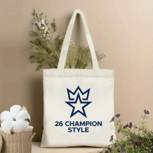 Front view of a beige canvas tote bag with a minimalist 2026 World Cup champion design. Features a sleek line-art logo combining a star and crown, paired with the bold "26 CHAMPION STYLE" text, styled with cotton stems and dried botanicals.
