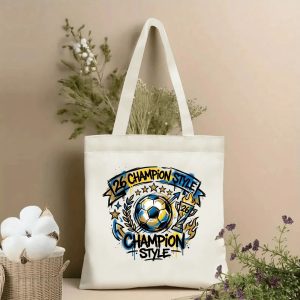 Front view of a beige canvas tote bag with a vibrant 2026 World Cup graffiti design. Features a central soccer ball, banner with "26 CHAMPION STYLE" lettering, stars, laurel wreath, trophy, flames, arrows, and street-art splatters, styled with cotton stems and dried botanicals.