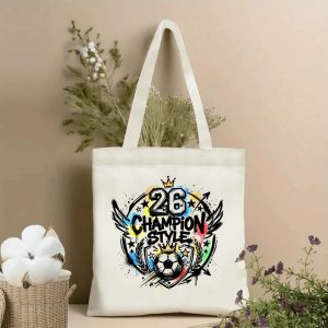 Front view of a beige canvas tote bag with a bold 2026 World Cup graffiti design. Features a central emblem with the number "26", "CHAMPION STYLE" lettering, a crown, soccer ball, wings, stars, laurel wreath, and dynamic street-art splatters, styled with cotton stems and dried botanicals.
