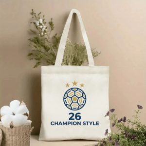 Front view of a beige canvas tote bag with a premium 2026 World Cup design. Features a stylized geometric soccer ball in deep navy blue with elegant gold accents, three shimmering gold stars above, and bold "26 CHAMPION STYLE" lettering in navy blue, styled with cotton stems and dried botanicals.