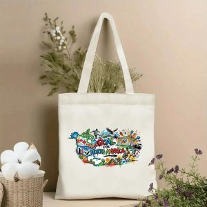 Front view of a beige canvas tote bag with a vibrant North America soccer design. Features a colorful graffiti-style map of North America, "GOAL FOR NORTH AMERICA" lettering, soccer players, a goal net, an eagle, cultural landmarks, and festive details, styled with cotton stems and dried botanicals.
