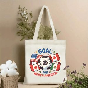 Front view of a beige canvas tote bag with a cute 2026 World Cup design. Features a smiling cartoon soccer ball, flags of the USA, Canada, and Mexico, and the "GOAL FOR NORTH AMERICA" slogan, styled with cotton stems and dried botanicals.