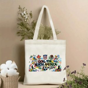 Front view of a beige canvas tote bag with a vibrant 2026 World Cup graffiti design. Features bold "GOAL FOR NORTH AMERICA" lettering, flags of Canada, USA, Mexico, a soccer ball, "WC 2026" text, and street-art style splatters, styled with cotton stems and dried botanicals.