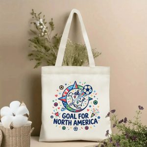 Angled shot of a canvas tote bag on a whitewashed wood surface, filled with magazines and daisies. Showcases the cheerful "Goal for North America" soccer compass graphic, perfect for game-day essentials.