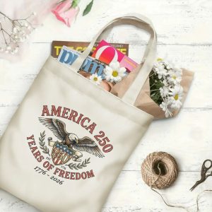Canvas tote bag with the same America 250th print, shown open with books and flowers inside, on a white wooden surface.
