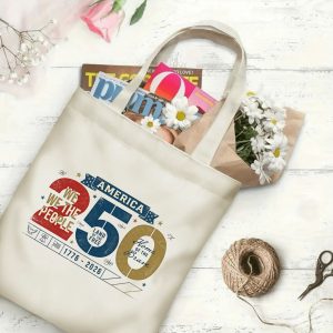 Canvas tote bag with the same America 250th print, shown open with books and flowers inside, on a white wooden surface.