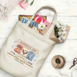 Canvas tote bag with the same America 250th print, shown open with books and flowers inside, on a white wooden surface.