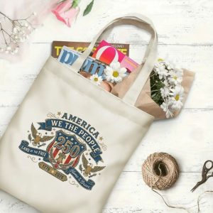 Canvas tote bag with the same America 250th crest print, shown open with books and flowers inside, on a white wooden surface.