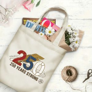 Canvas tote bag with the same America 250th print, shown open with books and flowers inside, on a white wooden surface.