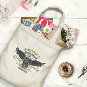 Canvas tote bag with the same America 250th print, shown open with books and flowers inside, on a white wooden surface.