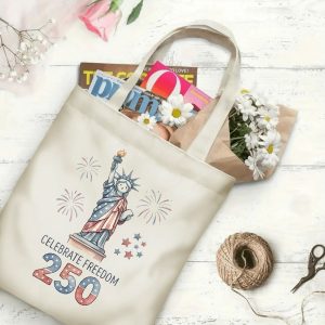 Canvas tote bag with the same America 250th print, shown open with books and flowers inside, on a white wooden surface.