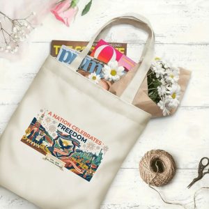 Canvas tote bag with the same America 250th print, shown open with books and flowers inside, on a white wooden surface.