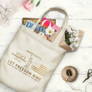 Canvas tote bag with the same America 250th print, shown open with books and flowers inside, on a white wooden surface.